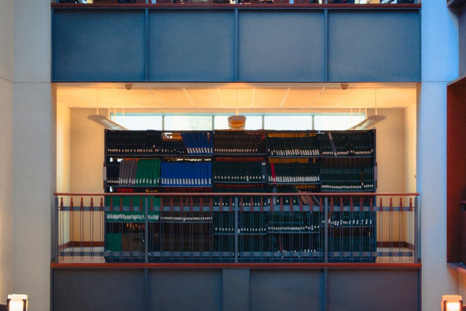The image shows a multi-tiered metal shelving unit filled with various coloured binders and files, positioned within an interior space overlooking a large window that allows natural light to illuminate the area. The shelves are situated on a small balcony or mezzanine level, which is enclosed by a thin orange metal safety railing. The binders are predominantly dark green, blue, and black, with some grey and red ones visible, and are made of plastic or cardboard with textured surfaces. The environment appears to be an office or storage area, with a ceiling featuring square acoustic tiles and white walls framing the scene. The lighting creates a warm glow around the shelves, contrasting with the bright daylight streaming through the window behind, partially obscured by the contents of the shelves. This setup relates to the storage of documents or office materials, illustrating a typical scenario where organised paper stock is maintained—an aspect possibly connected to private waste handling or archiving, linking indirectly to alternative rubbish management services such as those offered by Rubbish Collection Kingston.
