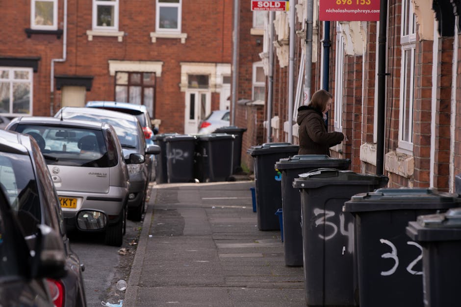 A woman standing on a residential pavement beside a row of black wheelie bins with closed lids, positioned in front of a brick terraced house with white-framed windows and a door. The bins are aligned along the edge of the sidewalk, some marked with white spray paint numbers. Several cars are parked on the street, close to the curb, with their rear and side windows visible. In the background, more terraced houses with similar brick facades and white detailing can be seen, along with additional bins placed against the buildings. Overhead, the sky appears overcast, casting diffuse natural light on the scene. The setting suggests an area where private waste collection services, such as those offered by Rubbish Collection Kingston, are used for street-side rubbish storage and collection, emphasizing the practicality of on-site waste management for residential properties in Kingston or similar urban environments.