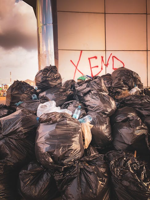 A large pile of black rubbish bags filled with waste materials is stacked against a modern tiled wall in an urban outdoor setting. The bags are made of glossy plastic and appear tightly packed, with some partially torn or unevenly sealed. Several clear plastic bottles are visible protruding from the bags, indicating common household waste. In the background, a graffiti tag with the letters “XEND” is spray-painted in red on the wall, and the sky above shows darker clouds, suggesting late afternoon or early evening lighting. The scene is located on a paved surface, possibly a sidewalk or outdoor area near a commercial or office building, with no additional waste containers or signage in view. This visual context reflects a private waste collection or on-site rubbish clearance activity, often managed by companies such as Rubbish Collection Kingston, to handle alternative waste disposal solutions outside of local authority services.