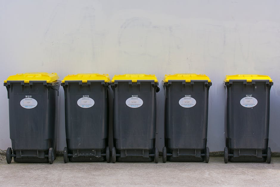 A row of five large wheeled rubbish bins positioned against a plain, light-colored wall on a concrete surface. The bins are made of dark grey plastic with textured surfaces and feature bright yellow lids, which are closed. Each bin has a white oval label with black text displaying identification numbers; the labels are centered on the front of each bin. The bins are evenly spaced and aligned in a straight horizontal line, with the wall behind them showing subtle shadows and a faint, uneven texture. The environment appears to be an outdoor area, possibly an alley or service yard, where rubbish collection or waste disposal occurs. The scene is well-lit with natural lighting, highlighting the smooth edges and surfaces of the bins, and emphasizing their utilitarian design. This image supports the concept of private waste handling and independent rubbish collection services offered by companies like Rubbish Collection Kingston, as visible in their typical setup for on-site waste management solutions.
