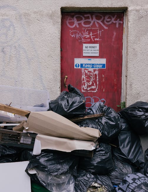 A cluttered scene in front of a weathered red door set into a beige, textured wall. The door is covered in graffiti and features a 'No Money' sign and a 'Keep Clear' sticker. In the foreground, there is a large pile of black rubbish bags, some partially torn, containing waste. Among the bags, there are flattened cardboard boxes, pieces of broken packaging, and discarded household items such as a wooden plank and what appears to be a white foam sheet. The waste is stacked on a paved surface, with small patches of greenery visible near the base of the wall. The overall environment suggests an urban area awaiting clearance or removal, emphasizing the need for professional rubbish disposal services like those offered by Rubbish Collection Kingston in handling private waste collection or on-site clearance projects.