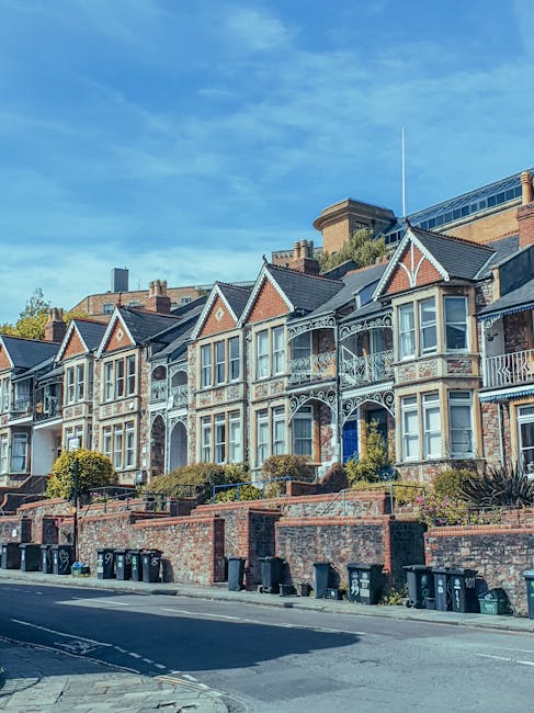 A row of Victorian-style terraced houses with decorative white ironwork on the balconies and window frames, built from red and brown brick with pitched roofs. The houses are situated on a raised pavement with small front gardens bordered by brick walls, some featuring bushes and small trees. In front of the houses, there is a street with black wheelie bins lined along the curb, indicating waste collection services. The sky above is bright blue with scattered light clouds, and a large modern building with glass windows is visible in the background behind the houses. The scene appears to be in a residential area where private waste collection, such as that offered by Rubbish Collection Kingston, may be involved in rubbish removal or house clearance processes, reflecting an environment where alternative waste handling methods are common.