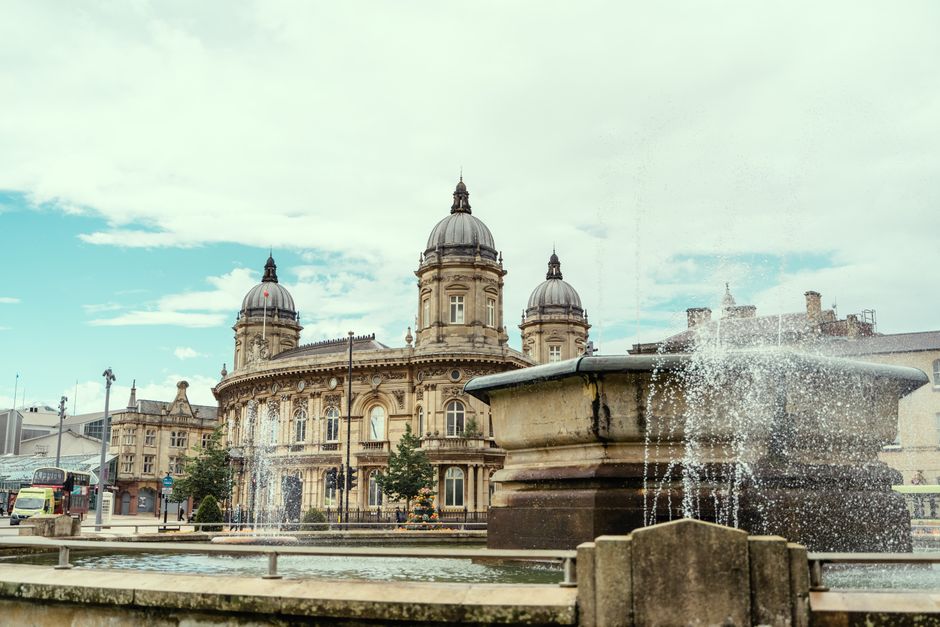 The image shows a historic sandstone building with multiple domed towers and ornate architectural details, situated behind a public fountain with water splashing from an elevated basin. The fountain, made of dark stone, has a wide, shallow bowl supported by a stepped base, with water cascading over the edges and creating visible spray. Towering above, the building features large arched windows, decorative stonework, and a central dome topped with a finial, all in warm beige and light brown tones with a weathered finish. The scene is set outdoors in an urban environment, with a paved open space or square in the foreground, and small trees lining the area. Light cloud cover and diffuse daylight illuminate the scene, with some shadows cast by the architectural elements. This image exemplifies the typical environment where independent waste collection, such as the kind provided by Rubbish Collection Kingston, might be employed for maintaining public spaces or handling waste disposal outside municipal services, aligning with private or alternative rubbish removal services.