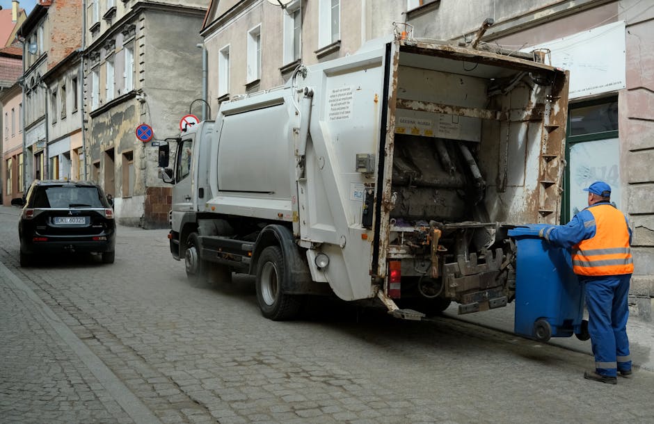 A white waste collection truck with a rusted and open rear compartment is parked on a narrow cobblestone street in an urban area, with a worker dressed in a blue uniform and an orange safety vest disposing of a blue recycling bin nearby. The truck's rear mechanism, used for emptying bins, is visible, showing mechanical components and a slightly worn, utilitarian finish. The surrounding environment includes multi-story buildings with a mix of stone and stucco facades, some with peeling paint, and small parked vehicles, including a black car further down the street. The scene is illuminated by natural daylight, casting soft shadows across the street, emphasizing the textures of the cobblestones, the truck's metal surfaces, and the worker's reflective clothing. This image illustrates the process of independent waste collection, a service offered as an alternative to local council rubbish removal, aligning with professional waste management practices provided by companies like Rubbish Collection Kingston involved in rubbish services.