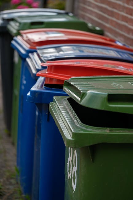 A row of four large waste collection bins positioned along a pavement next to a brick wall, with the closest bin being green and featuring a ridged lid, followed by a blue bin with a smooth lid, a red bin with a smooth lid, and another green bin with a ridged lid further back. The bins are made of durable plastic with textured surfaces, and they are arranged side-by-side in an outdoor setting, likely used for domestic rubbish collection. The pavement surface appears to be concrete, and the background shows a blurred view of other bins and part of a residential or commercial building. The scene is lit with natural daylight, highlighting the vivid colours and clean condition of the bins, which are sometimes used by private waste handling services like Rubbish Collection Kingston, potentially serving as an alternative to council-managed rubbish disposal. The image underscores organized waste collection in an urban environment, suitable for contexts involving on-site clearance or private rubbish collection services.