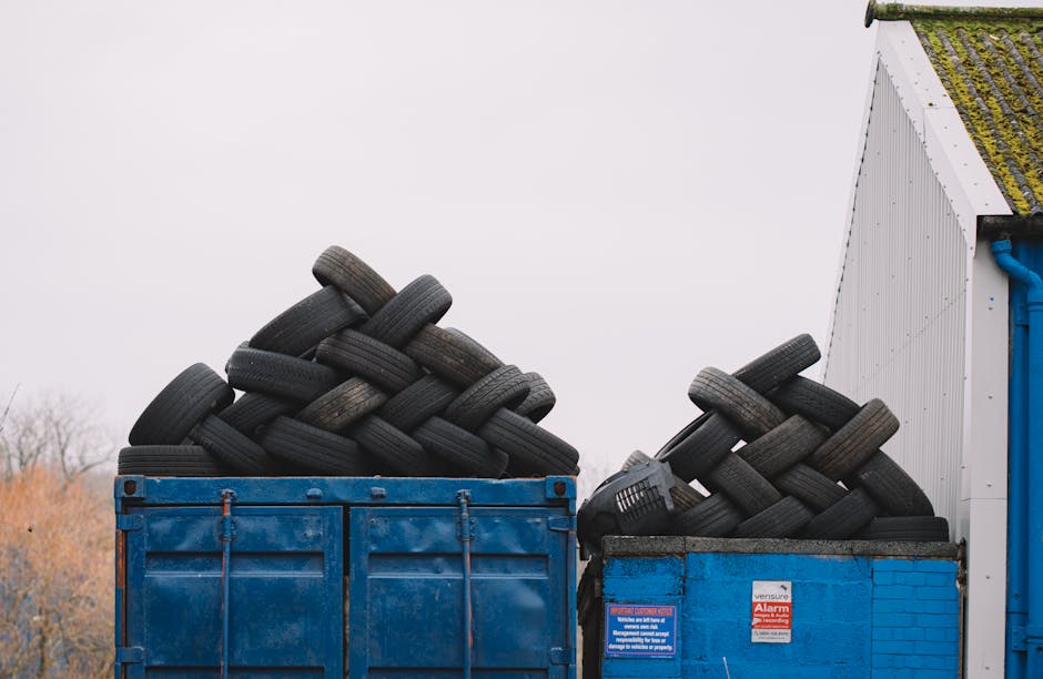 Two large open-top containers filled with discarded black rubber tires are situated outdoors, with the container on the left being a blue metal skip and the one on the right constructed of blue brickwork with a black top. The tires are stacked irregularly, with some leaning and overlapping, displaying visible treads and sidewalls with a weathered appearance. The rubber tires vary in size and shape, some with flat profiles and others with slight bulges, and show signs of dirt and wear. The containers are positioned adjacent to a white building with a barcode and warning labels, and are partially covered by overhanging eaves that have moss and dirt on their surfaces. The environment appears to be an industrial or waste management site, with a background of leafless trees and an overcast sky, suggesting a cool or damp day. The scene reflects a private approach to waste collection, supporting independent rubbish removal services such as those offered by Rubbish Collection Kingston.