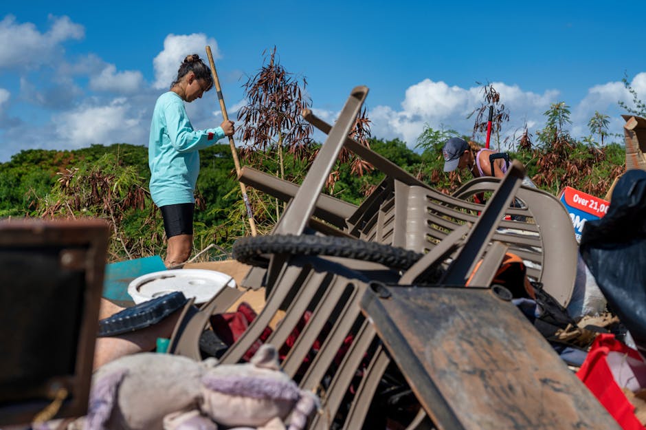 The image depicts two individuals working outdoors amidst a large accumulation of discarded items and rubbish, which includes plastic chairs, a white circular plastic tray, a wooden table, and various other forms of debris. The person on the left, dressed in a light blue long-sleeve top and black shorts, is holding a long-handled tool and appears to be sorting or lifting waste. The individual on the right, wearing a sleeveless top, a cap, and sunglasses, is partially crouched and engaged in clearing or arranging the rubbish pile. The surrounding environment features a clear blue sky with a few white clouds, lush green trees and shrubs, and some brown dead or leafless branches visible behind the waste. The scene is illuminated by natural daylight, casting minimal shadows, highlighting the textures of the plastic, wood, and metal debris. This setting illustrates an informal waste collection or clearance effort, reflective of private or on-site rubbish handling, which relates to services provided by companies such as Rubbish Collection Kingston for efficient rubbish removal and disposal.