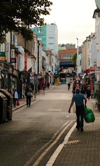 A busy urban street scene featuring a mix of low-rise and high-rise buildings lining both sides of the road, with pedestrians walking along the sidewalk and crossing the street. In the foreground, a man wearing a teal shirt is walking away from the camera, carrying a large green bag, while other people stroll or stand near shopfronts and entrances. The buildings display a variety of architectural styles and colors, with some storefront signage visible. The street appears to be temporarily closed to traffic to accommodate foot traffic, and there is a gentle incline leading towards a modern glass-fronted building at the far end. Mature trees provide some greenery and shade along the sidewalk. The overall setting suggests an active commercial area, where private waste collection services such as those from Rubbish Collection Kingston might operate, especially in context of alternative rubbish removal and on-site clearance activities nearby, evidenced by the arrangement of the street and the mixed-use environment.