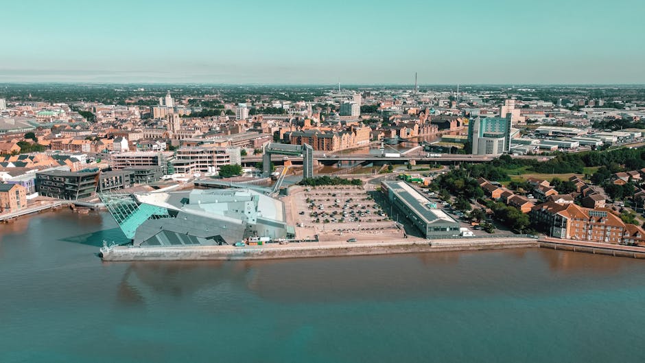 An aerial view of a cityscape showing a large body of water in the foreground, with an industrial or commercial complex situated along the shoreline. The complex includes a modern, teal-colored building with angular, geometric roofing, surrounded by paved areas and parked vehicles, indicating a loading or storage zone. Adjacent to the building are several other structures made from concrete and metal, with flat roofs and large windows, all set within a partly landscaped area with trees and greenery. In the background, there are numerous mid-rise and high-rise buildings, including office blocks, residential apartments, and possibly a few landmarks, extending into the distance under a clear, green-tinged sky, suggesting bright daylight. This scene reflects urban development alongside water, emphasizing elements typical of private waste handling and independent rubbish collection services, such as Rubbish Collection Kingston, operating in the central Kingston area.