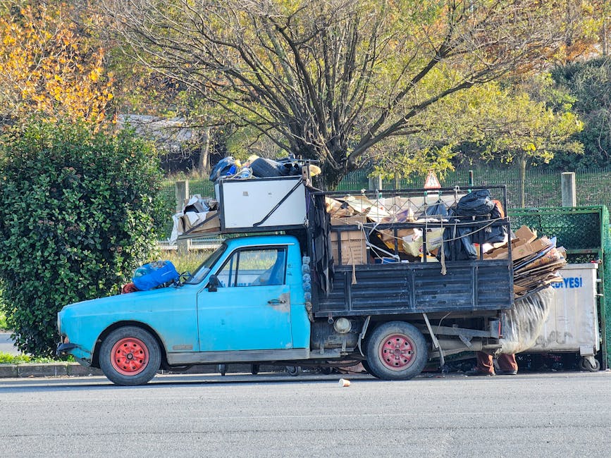 A small, older blue pickup truck parked parallel to the curb on a city street, heavily loaded with various types of rubbish including large cardboard boxes, black plastic bags, and miscellaneous debris piled high past the truck’s sides. The truck’s body shows signs of wear with faded paint and minor dents, and its red-painted wheels are visible beneath the load. The workload extends into the truck’s black metal cargo bed, which is surrounded by a simple safety rail, with some items partially secured by ropes. In the background, there is a tall tree with green and yellow leaves indicating early autumn, and a brown fence behind a grassy area. The scene is illuminated by natural daylight, emphasizing the cluttered nature of this on-site waste accumulation, which aligns with alternative waste handling or private rubbish collection services, such as those offered by Rubbish Collection Kingston, outside of regular municipal rubbish pickup. The overall setting reflects a typical street scene where private or independent rubbish clearance might be taking place, highlighting the necessity of reliable rubbish removal services for efficient waste disposal.