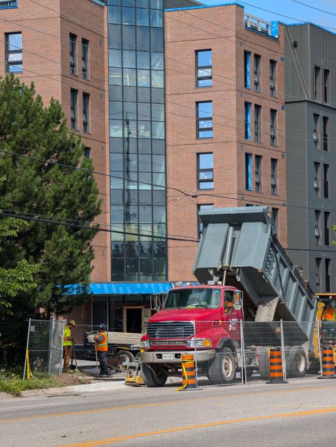 A red skip truck with a raised metal container is parked on the street in front of a modern multi-storey building with a brick facade and glass windows. The truck is positioned behind a temporary metal fence with orange and black striped safety barriers placed around it. Several workers wearing high-visibility vests and helmets are seen near the construction site, engaged in activities related to waste removal or construction. The scene is illuminated by daylight, with a large leafy tree partially visible on the left side, and overhead power lines crossing the image. The setting appears to be an urban area, possibly undergoing building renovations or extensions, highlighting private or independent waste collection services involved in managing construction debris amid ongoing construction delays or alternative rubbish handling processes.